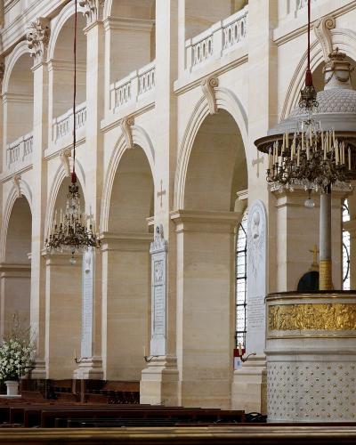 The interior of a grand stone church featuring a prominent marble and gold pulpit, arched colonnades, hanging chandeliers, and French flags.