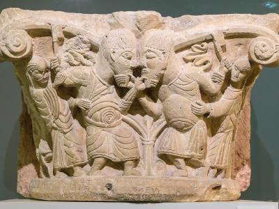 A close-up photograph of an ancient stone relief sculpture, likely a capital or architectural fragment, featuring carved figures in classical attire and inscriptions at the base, displayed on a white pedestal against a muted green wall.