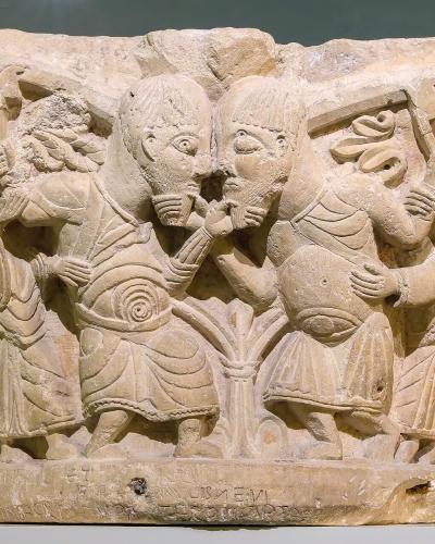 A close-up photograph of an ancient stone relief sculpture, likely a capital or architectural fragment, featuring carved figures in classical attire and inscriptions at the base, displayed on a white pedestal against a muted green wall.