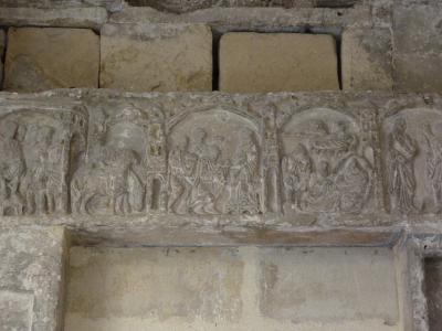 A close-up photograph of a weathered stone architectural frieze featuring carved relief sculptures within arched niches, set into an aged masonry wall.