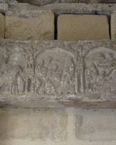 A close-up photograph of a weathered stone architectural frieze featuring carved relief sculptures within arched niches, set into an aged masonry wall.