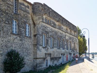 A long, historic stone building with arched windows and a fortified corner tower, situated along a riverside walkway with a distinctive curved lamppost and a person walking nearby.