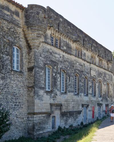 A long, historic stone building with arched windows and a fortified corner tower, situated along a riverside walkway with a distinctive curved lamppost and a person walking nearby.
