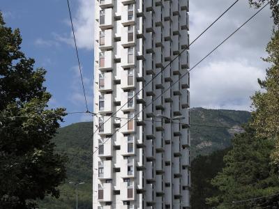 A tall, white, brutalist-style residential tower with a distinctive geometric facade of protruding balconies and windows, situated on a street with cars, trees, and hills in the background under a partly cloudy sky.
