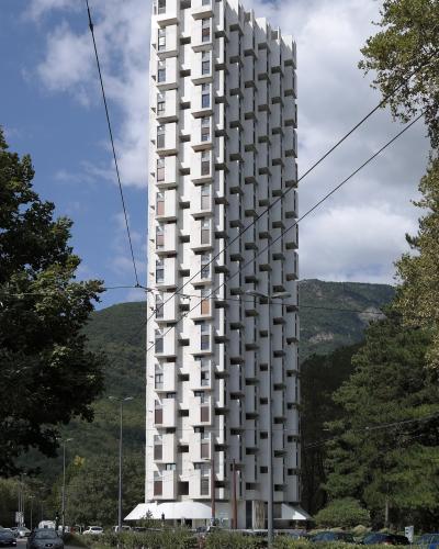 A tall, white, brutalist-style residential tower with a distinctive geometric facade of protruding balconies and windows, situated on a street with cars, trees, and hills in the background under a partly cloudy sky.
