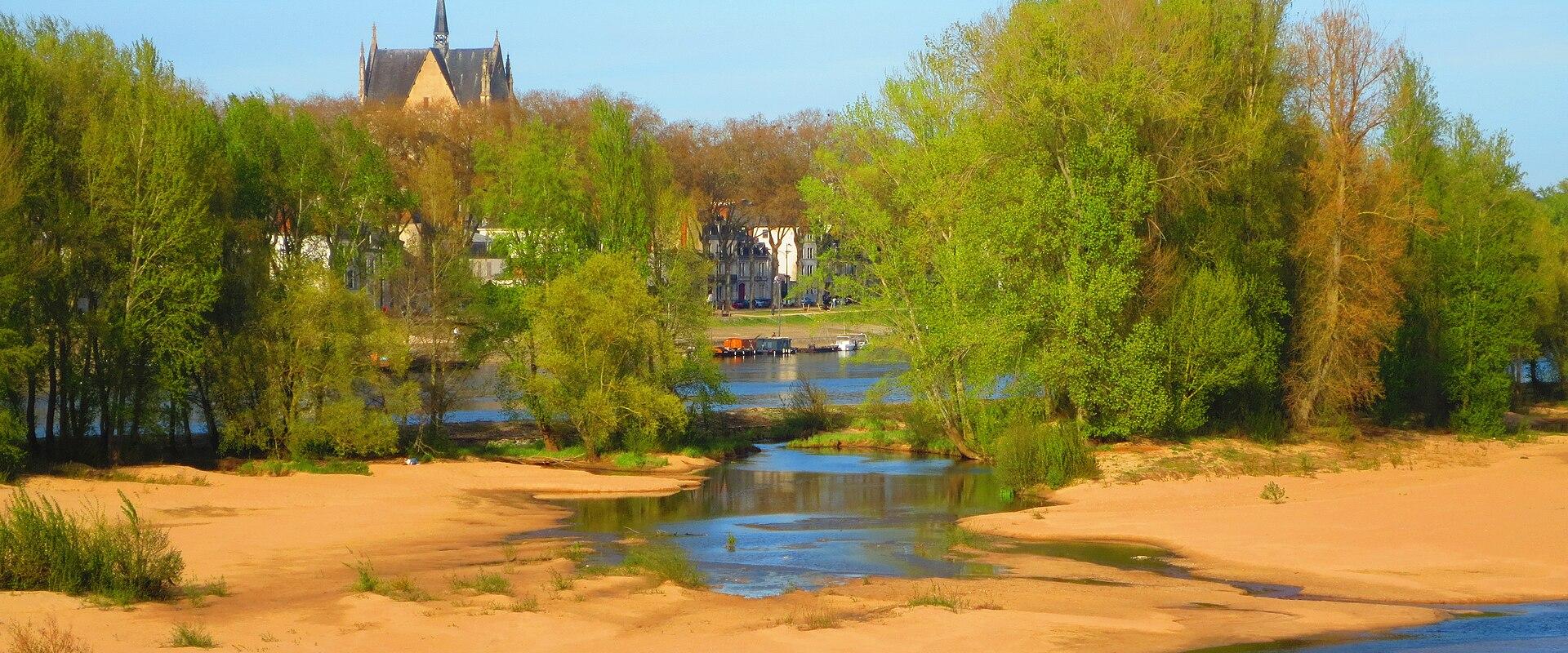 A riverside scene with sandy banks, green trees, a small stream inlet, and a Gothic-style church spire visible in the background under a clear blue sky.