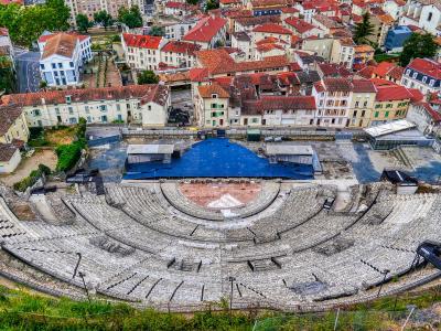 A high-angle view of the Roman Theatre of Vienne showing its semi-circular stone seating tiers, a blue-covered stage area, and surrounding historic buildings with red-tiled roofs.