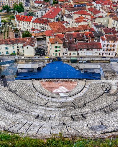 A high-angle view of the Roman Theatre of Vienne showing its semi-circular stone seating tiers, a blue-covered stage area, and surrounding historic buildings with red-tiled roofs.