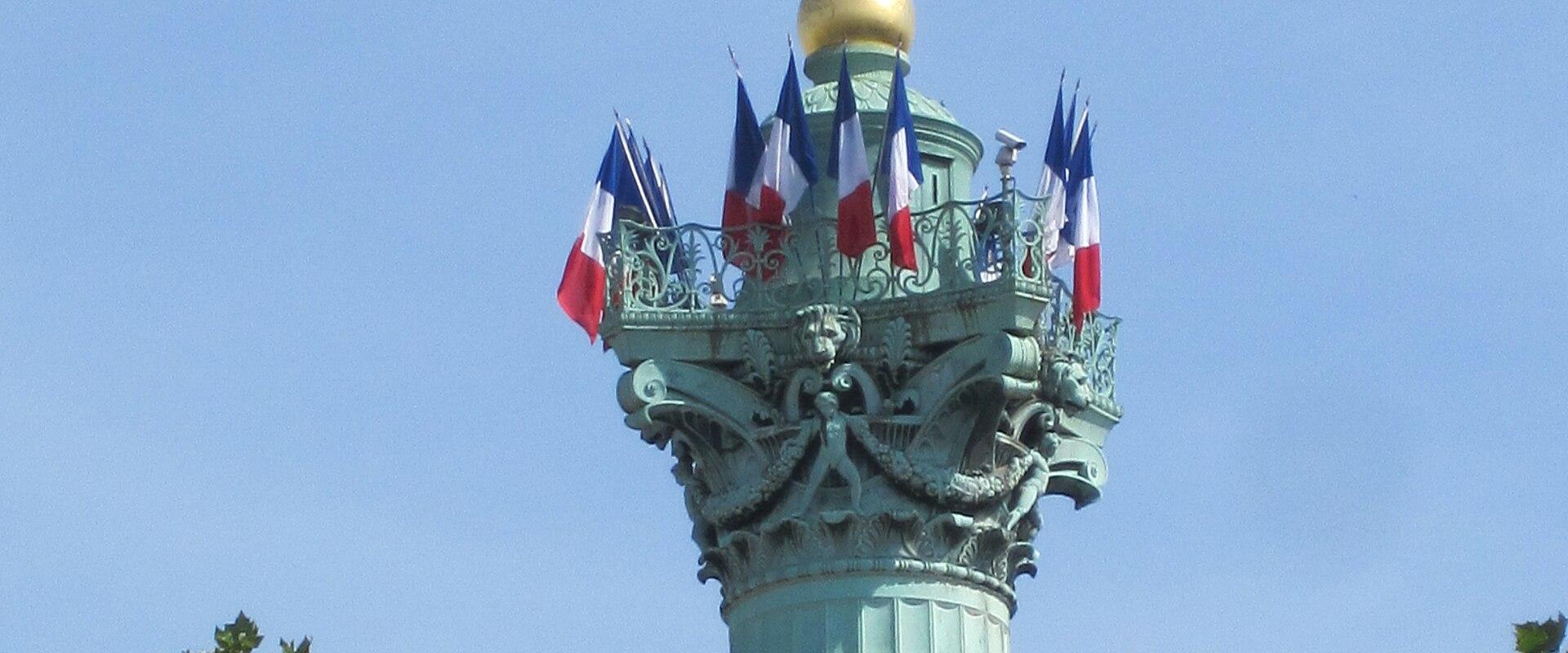 The image shows the top of the July Column (Colonne de Juillet) in Place de la Bastille, Paris, featuring a golden statue of the Spirit of Liberty atop a fluted column decorated with French flags and ornate bronze reliefs, framed by green tree fol