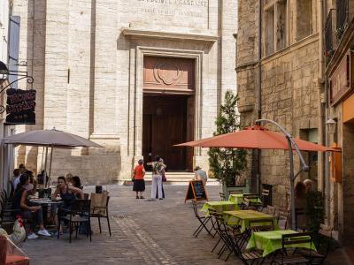 The image shows the stone facade of a large church or cathedral with a prominent wooden door, an inscription reading "REPUBLIQUE FRANCAISE" above the entrance, and outdoor café seating on a cobblestone street in front.