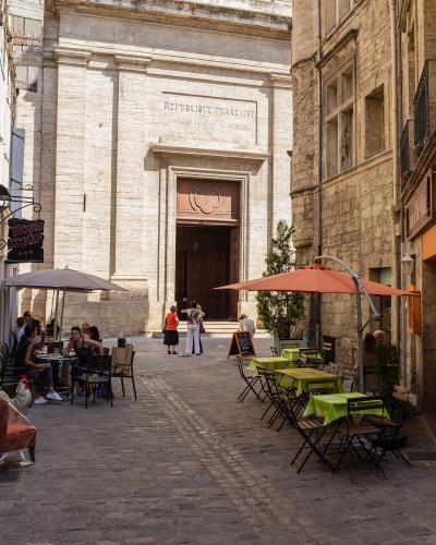 The image shows the stone facade of a large church or cathedral with a prominent wooden door, an inscription reading "REPUBLIQUE FRANCAISE" above the entrance, and outdoor café seating on a cobblestone street in front.
