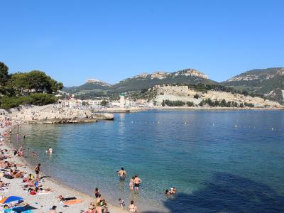 A crowded pebble beach with people sunbathing and swimming in clear blue water, backed by a stone seawall, a harbor with a lighthouse, and a hilltop fortress (Château de Cassis) under a clear sky.
