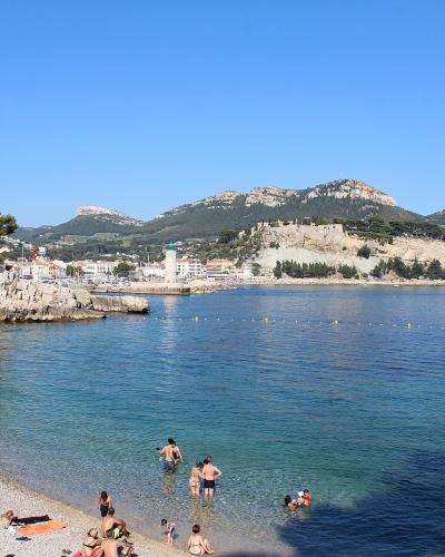 A crowded pebble beach with people sunbathing and swimming in clear blue water, backed by a stone seawall, a harbor with a lighthouse, and a hilltop fortress (Château de Cassis) under a clear sky.