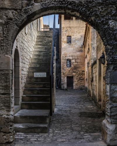 A narrow cobblestone alleyway in a historic stone village, viewed through a rough-hewn stone archway, with an external stone staircase and a sign reading "Privé - Accès interdit / Private - No access".