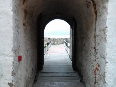 A view through a whitewashed stone archway with exposed brick voussoirs, leading onto a wooden boardwalk that exits to an outdoor terrace with railings overlooking water.