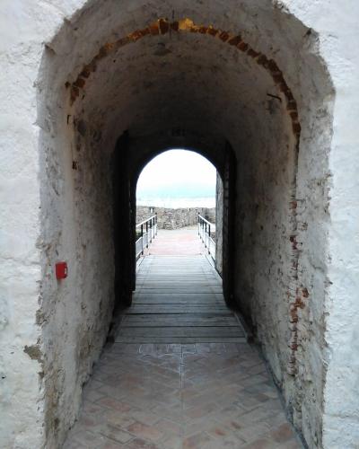 A view through a whitewashed stone archway with exposed brick voussoirs, leading onto a wooden boardwalk that exits to an outdoor terrace with railings overlooking water.