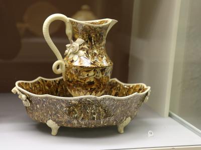 A ceramic pitcher and basin set with a mottled brown-and-cream glaze and white relief floral decorations, displayed inside a glass museum case on a white shelf with a small label "12".