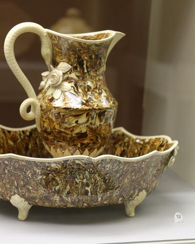 A ceramic pitcher and basin set with a mottled brown-and-cream glaze and white relief floral decorations, displayed inside a glass museum case on a white shelf with a small label "12".