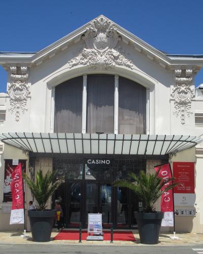 A white, ornate building facade with a central entrance marked "CASINO" in white letters above the doors, flanked by red banners and potted palm trees under a clear blue sky.