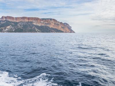 A wide seascape showing a massive, flat-topped coastal cliff with reddish-brown and ochre rock layers rising steeply from the sea, viewed from a moving boat with white wake in the foreground.