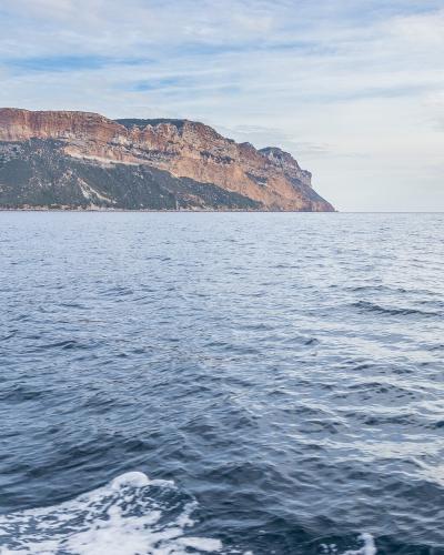 A wide seascape showing a massive, flat-topped coastal cliff with reddish-brown and ochre rock layers rising steeply from the sea, viewed from a moving boat with white wake in the foreground.
