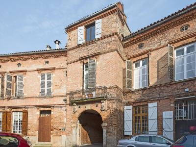The image shows a multi-story brick building with an arched passageway, shuttered windows, and a small balcony, with a red car and a silver car parked in front under a blue sky.