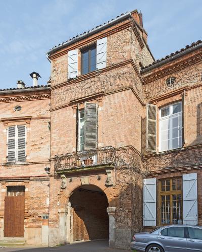 The image shows a multi-story brick building with an arched passageway, shuttered windows, and a small balcony, with a red car and a silver car parked in front under a blue sky.