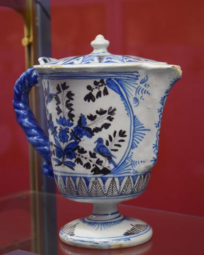 A close-up photograph of a white ceramic pot with blue and black floral and bird motifs, displayed on a glass shelf against a red background, with a museum label in the foreground reading "Pot de pharmacie couvert".
