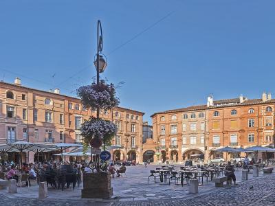 Cobblestone square with red brick buildings, arcades, and a central lamp post under a blue sky.