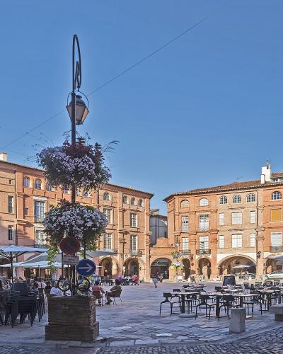 Cobblestone square with red brick buildings, arcades, and a central lamp post under a blue sky.