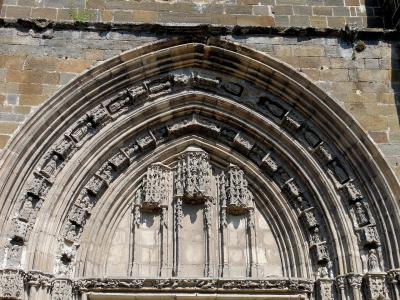 A close-up photograph of an ornate Gothic stone portal featuring multiple concentric arches (archivolt) with sculpted figures and intricate tracery, set within a weathered stone wall.