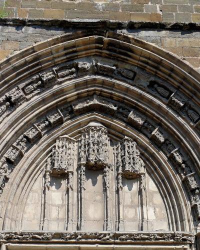 A close-up photograph of an ornate Gothic stone portal featuring multiple concentric arches (archivolt) with sculpted figures and intricate tracery, set within a weathered stone wall.