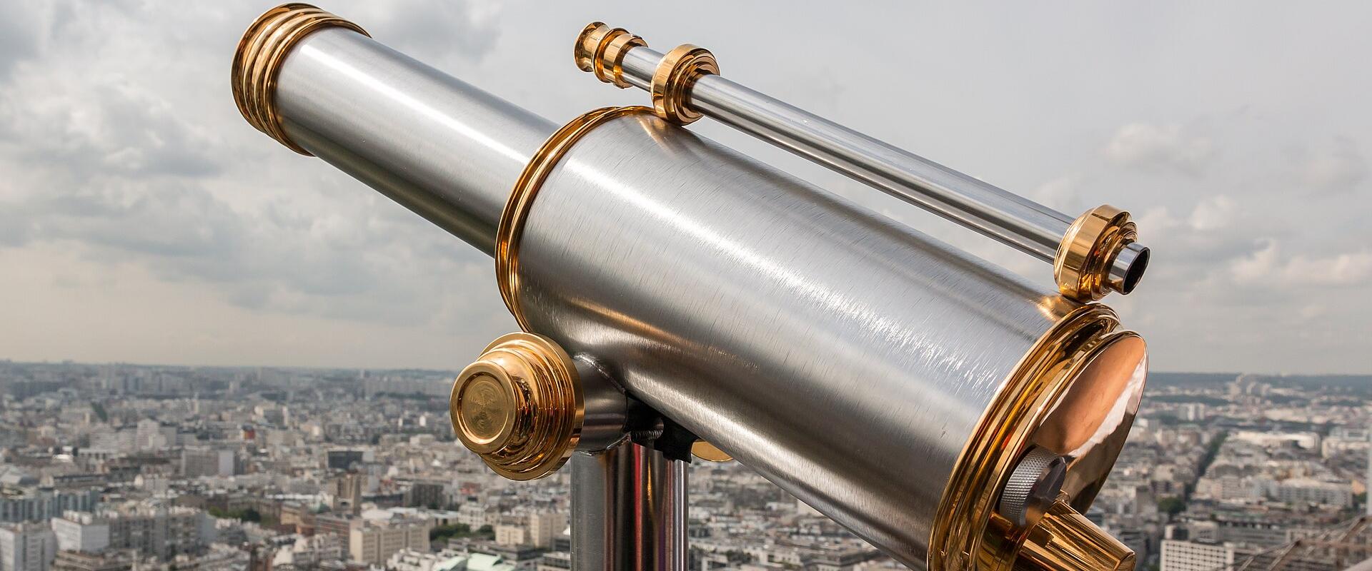 A close-up of a silver and gold coin-operated telescope on a viewing platform, overlooking a dense urban cityscape with uniform Haussmann-style buildings and green spaces.