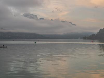 A wide shot of a calm lake at dusk or dawn with misty mountains in the background, featuring a small wooden dock on the left, red and green navigation buoys, and a hazy sky.