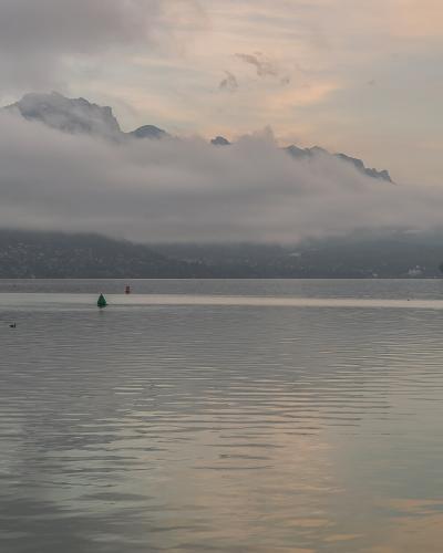 A wide shot of a calm lake at dusk or dawn with misty mountains in the background, featuring a small wooden dock on the left, red and green navigation buoys, and a hazy sky.