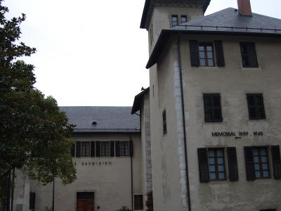 The image shows the exterior of a historic building complex with a tower, featuring signage that reads "LE SAVOISIEN" on one wing and "MEMORIAL 1939 - 1945" on another, set within a landscaped courtyard with benches and flowerbeds.