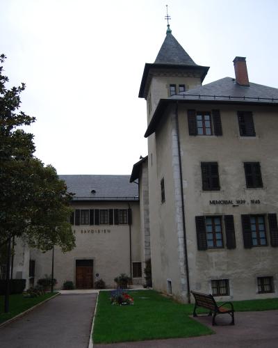 The image shows the exterior of a historic building complex with a tower, featuring signage that reads "LE SAVOISIEN" on one wing and "MEMORIAL 1939 - 1945" on another, set within a landscaped courtyard with benches and flowerbeds.