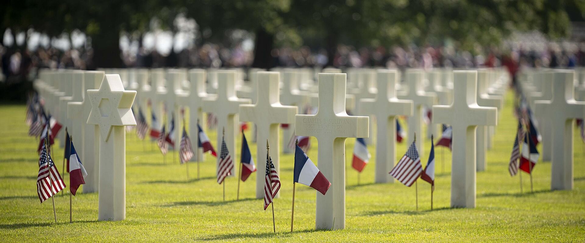 1. Rows of white marble headstones (Latin crosses and Star of David) in a manicured green lawn, each marked with small American and French flags, with blurred trees and spectators in the background.