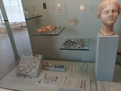 A museum display case containing small Gallo-Roman artifacts including pottery, jewelry, pins, and a terracotta bust on a pedestal.