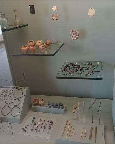 A museum display case containing small Gallo-Roman artifacts including pottery, jewelry, pins, and a terracotta bust on a pedestal.