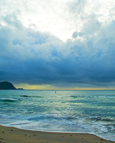 Dramatic cloudy sky over turquoise ocean waves breaking on a sandy beach with a distant headland.