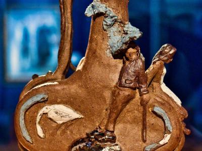 A close-up photograph of a brown ceramic pilgrim flask (gourde) with sculpted figures and decorative elements, displayed on a stand in a museum setting with blue background lighting.