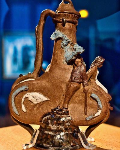 A close-up photograph of a brown ceramic pilgrim flask (gourde) with sculpted figures and decorative elements, displayed on a stand in a museum setting with blue background lighting.