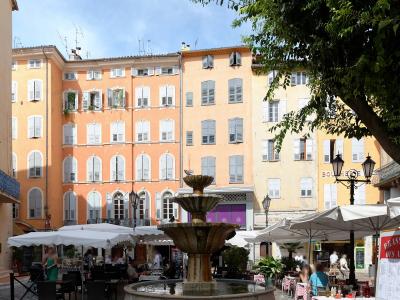 A sunlit public square in Grasse, France, featuring a central stone fountain, surrounding café tables with red-and-white checkered tablecloths, and pastel-colored buildings with white shutters; signage includes "BOULANGERIE" and a menu board readi