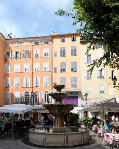 A sunlit public square in Grasse, France, featuring a central stone fountain, surrounding café tables with red-and-white checkered tablecloths, and pastel-colored buildings with white shutters; signage includes "BOULANGERIE" and a menu board readi