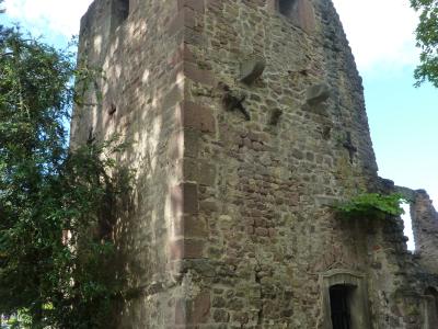 A low-angle photograph of a weathered, medieval stone tower with arched openings near the top, set against a blue sky with trees on the left.