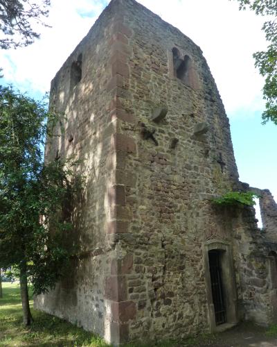 A low-angle photograph of a weathered, medieval stone tower with arched openings near the top, set against a blue sky with trees on the left.