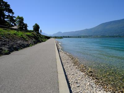 A paved promenade runs alongside a pebble beach and turquoise water, with mountains in the background under a clear blue sky.