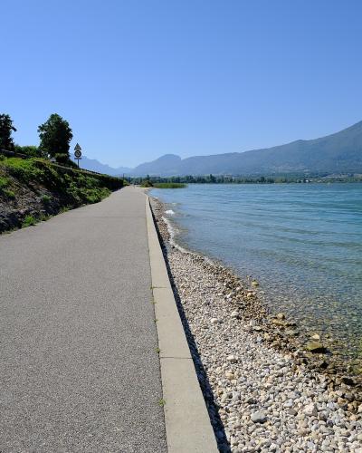 A paved promenade runs alongside a pebble beach and turquoise water, with mountains in the background under a clear blue sky.