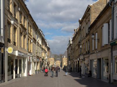 A pedestrianized urban street lined with multi-story stone buildings housing retail shops, featuring overhead tram or trolleybus wires and a distant classical building at the terminus.